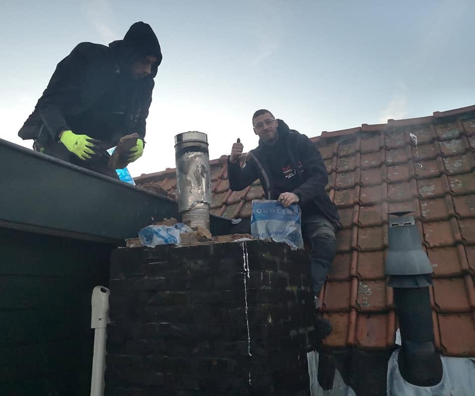 De dakdekkers van Moonen op een pannendak bij een schoorsteen, een van hen steekt zijn duim op