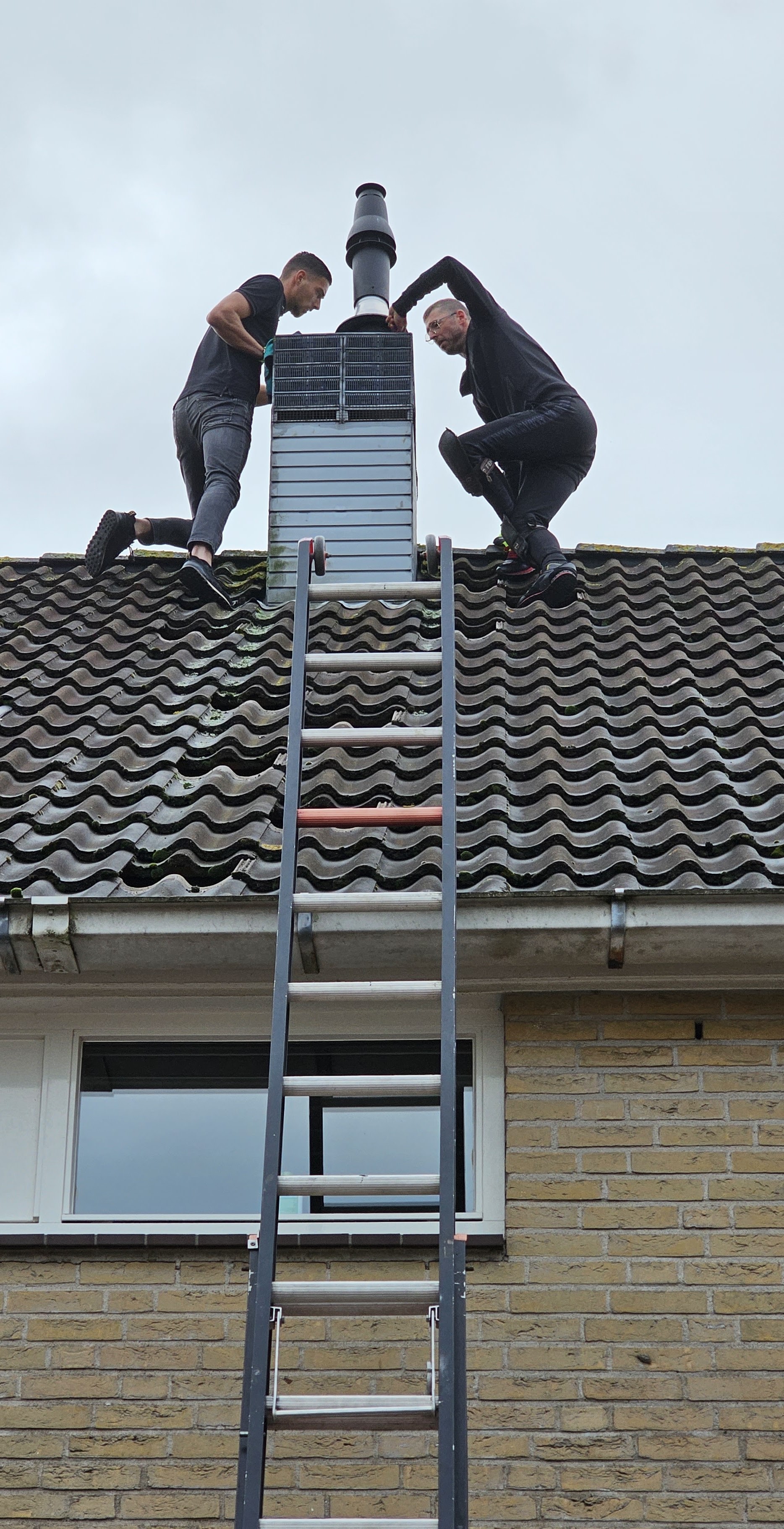 Twee dakdekkers aan het werk bij een rookkanaal op een pannendak, met een ladder tegen de gevel